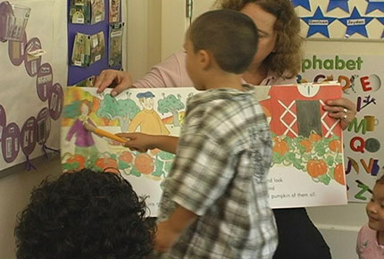 A child uses a pointer to point to an illustration in a book held by his teacher