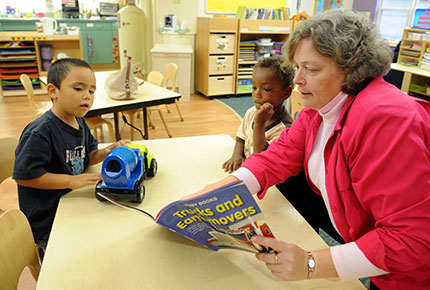 An adult reads a book about construction vehicles to two preschoolers sitting at a table. One boy holds a toy cement mixer