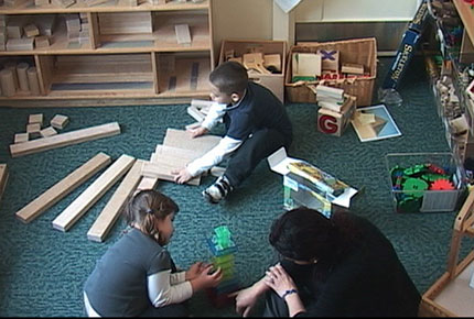 A preschool teacher sits on the floor to engage a child stacking geometric shapes. Another child plays with large blocks nearby.