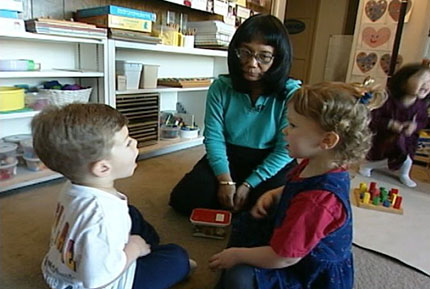 A preschool teacher kneels on the floor with two preschoolers, who are facing each other