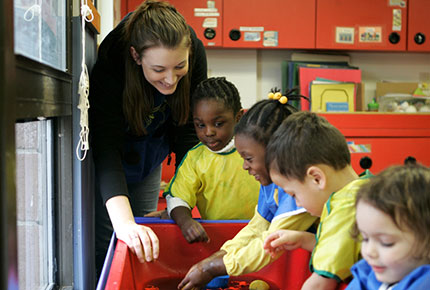 A preschool teacher smiles as she engages four preschoolers at the water table