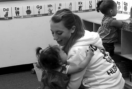 A caregiver smiles down at an older infant, who has wrapped her arms around the caregiver