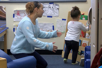 A teacher guides a toddler to put a book away on the display shelf