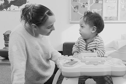 An infant stands at an activity table and looks at her teacher, who is smiling at the infant