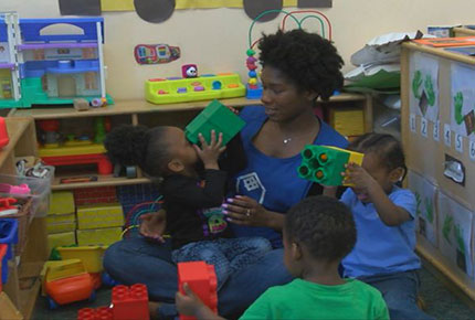 A teacher sits on the floor with three children building with large blocks. 