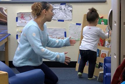 A toddler puts away a book as her teacher guides her