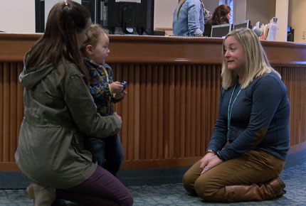 A teacher kneels on the floor, talking to a toddler visiting with her mother