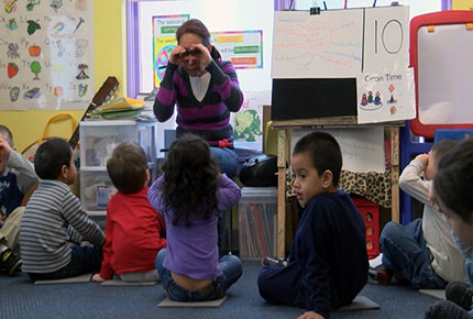 A teacher holds her hands up to her eyes like binoculars while sitting in front of a large group of children.