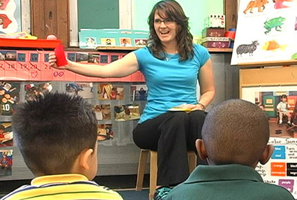 A teacher sits in front of a group of students and is holding something up for them to see.