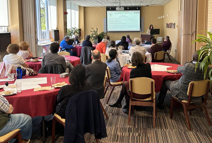 Presenter at podium with slides speaking to a room full of faculty at round tables