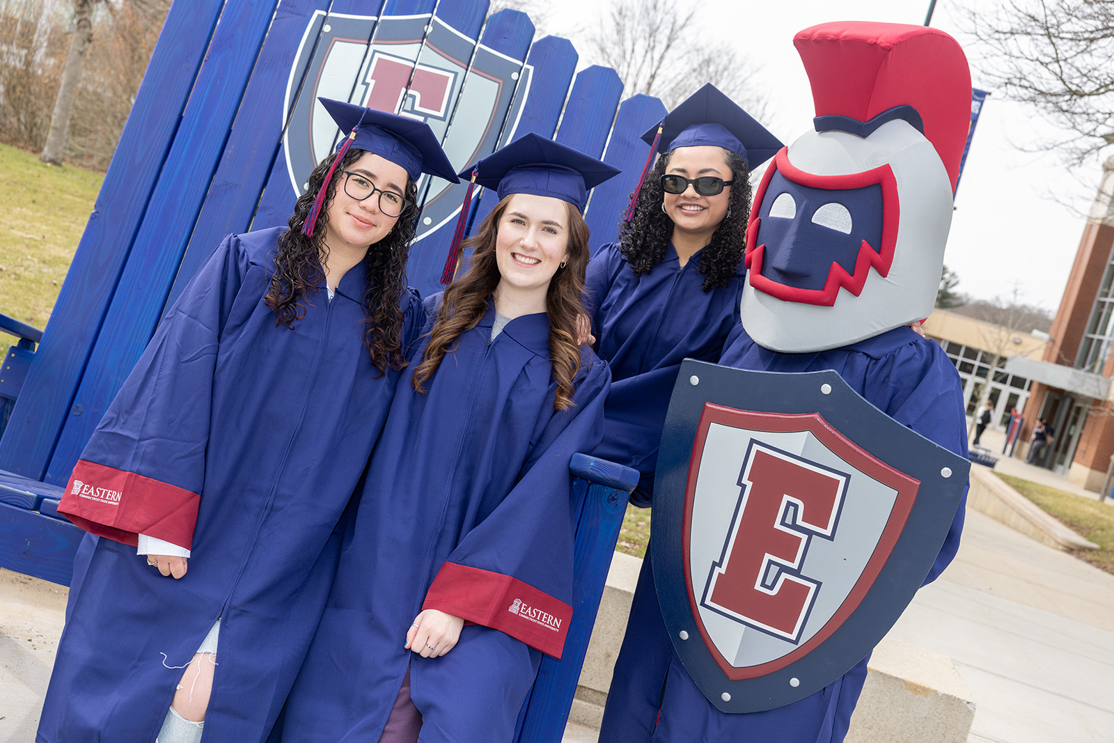 Students wearing cap and gowns with Willi the Warrior in front of a large blue chair with the Eastern shield on it