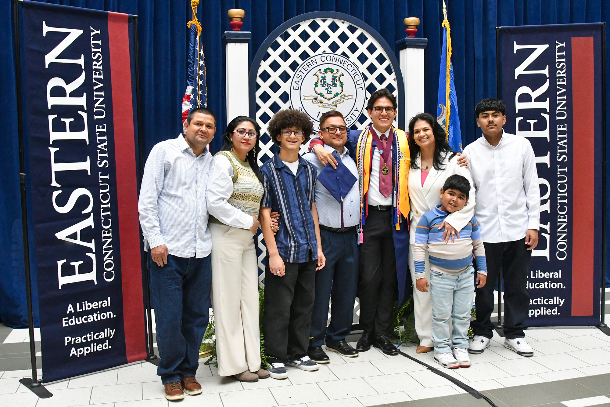 Graduate in gown holding cap posing for photo with family members in front of a blue backdrop with the Eastern crest, American and Connecticut state flag, and vertical Eastern banners on the side