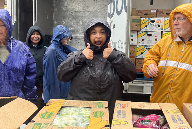 student giving thumbs up while wearing rain gear and unpacking boxes of food from a truck