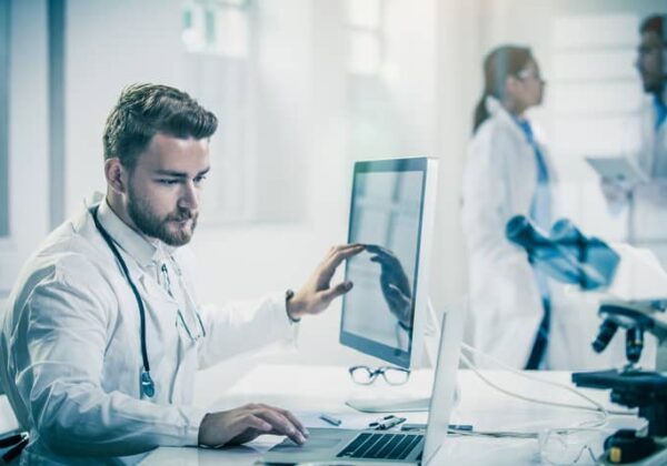 Male healthcare professional comparing data on a desktop vs. laptop.