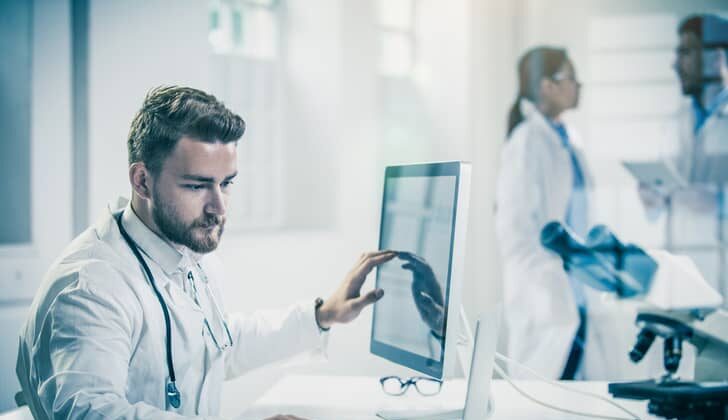 Male healthcare professional comparing data on a desktop vs. laptop.