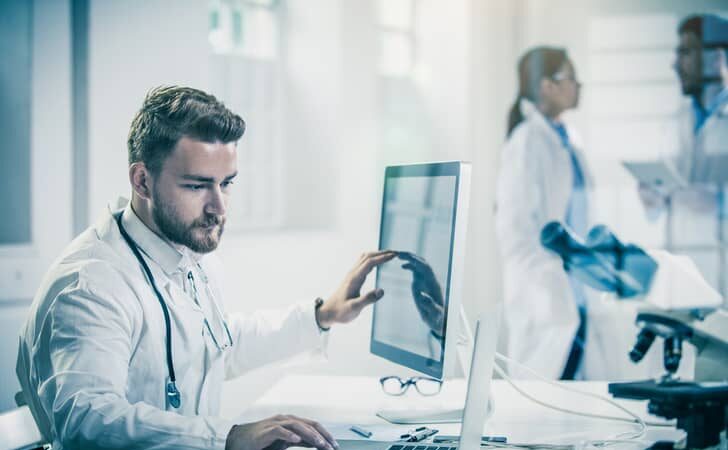 Male healthcare professional comparing data on a desktop vs. laptop.