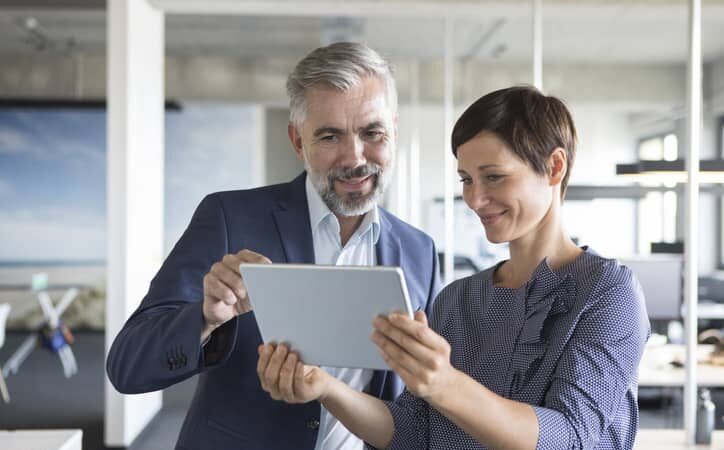 A businessman and businesswoman look together at a tablet in an office.