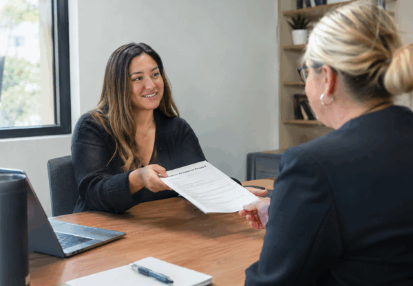 A woman meets with an admissions or academic advisor in an office, smiling as she hands over a document across a desk with a laptop and notepad nearby.