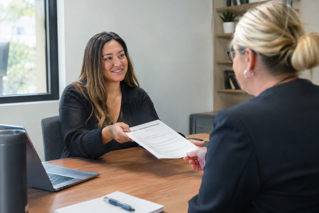 A woman meets with an admissions or academic advisor in an office, smiling as she hands over a document across a desk with a laptop and notepad nearby.