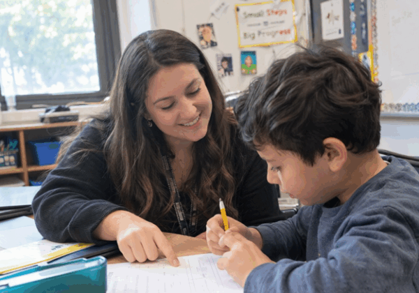 A teacher points to a paper as a young student works on an assignment, providing individual guidance and support in a learning environment.