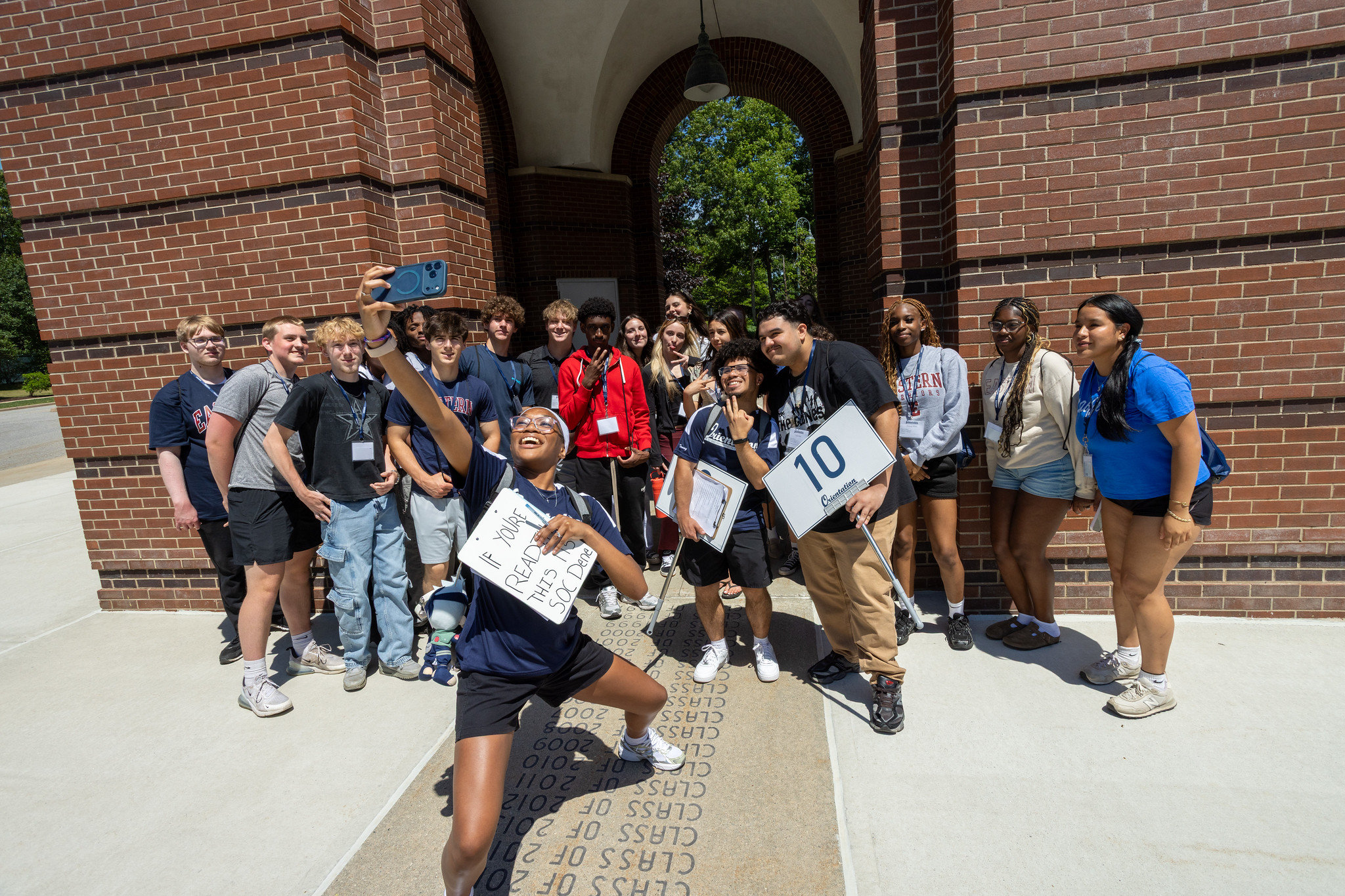 An orientation leader taking a selfie with a group of new students under the campus archway