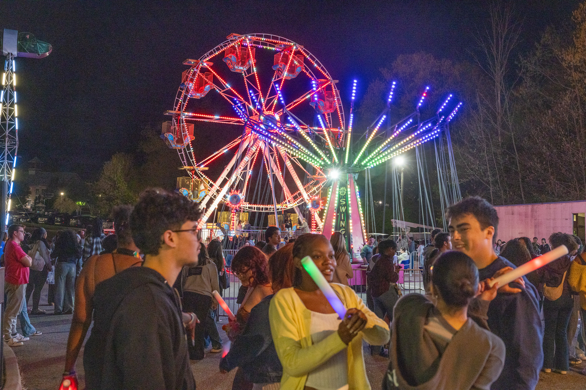 Students with glow sticks at a nighttime campus carnival with a lit Ferris wheel
